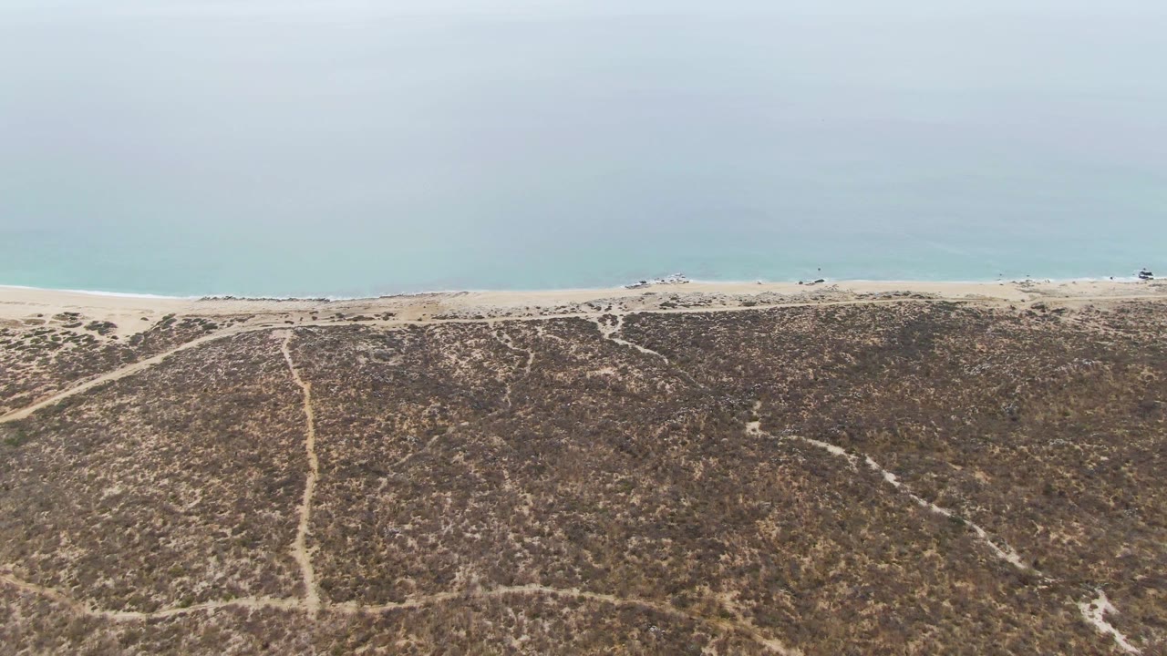 Aerial frame at Punta Gorda Beach showing branching tracks across dry dune vegetation, a narrow sand strip, small vehicles near the shore, blue-green ocean, and no visible people.