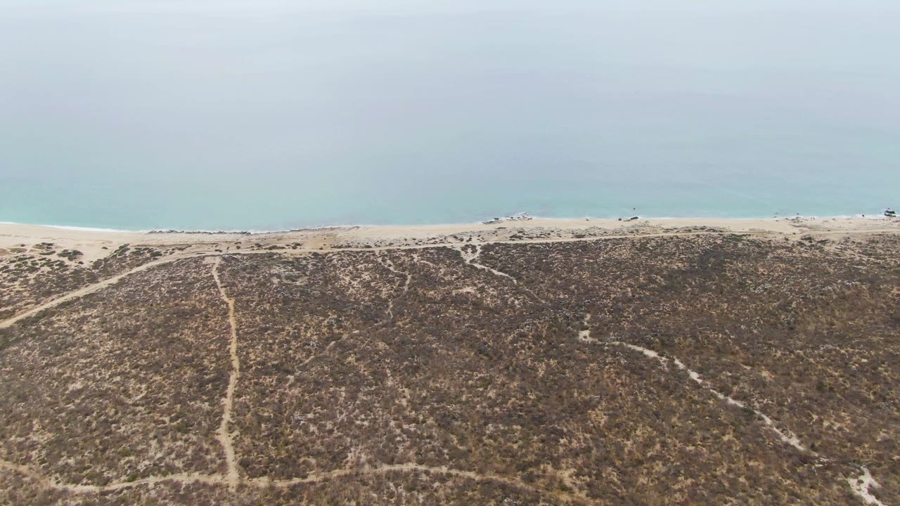 Mid-clip aerial view of Punta Gorda Beach with dry textured dunes filling the lower frame, pale tire tracks cutting toward the shore, several small vehicles on the sand, calm water, and no visible people.
