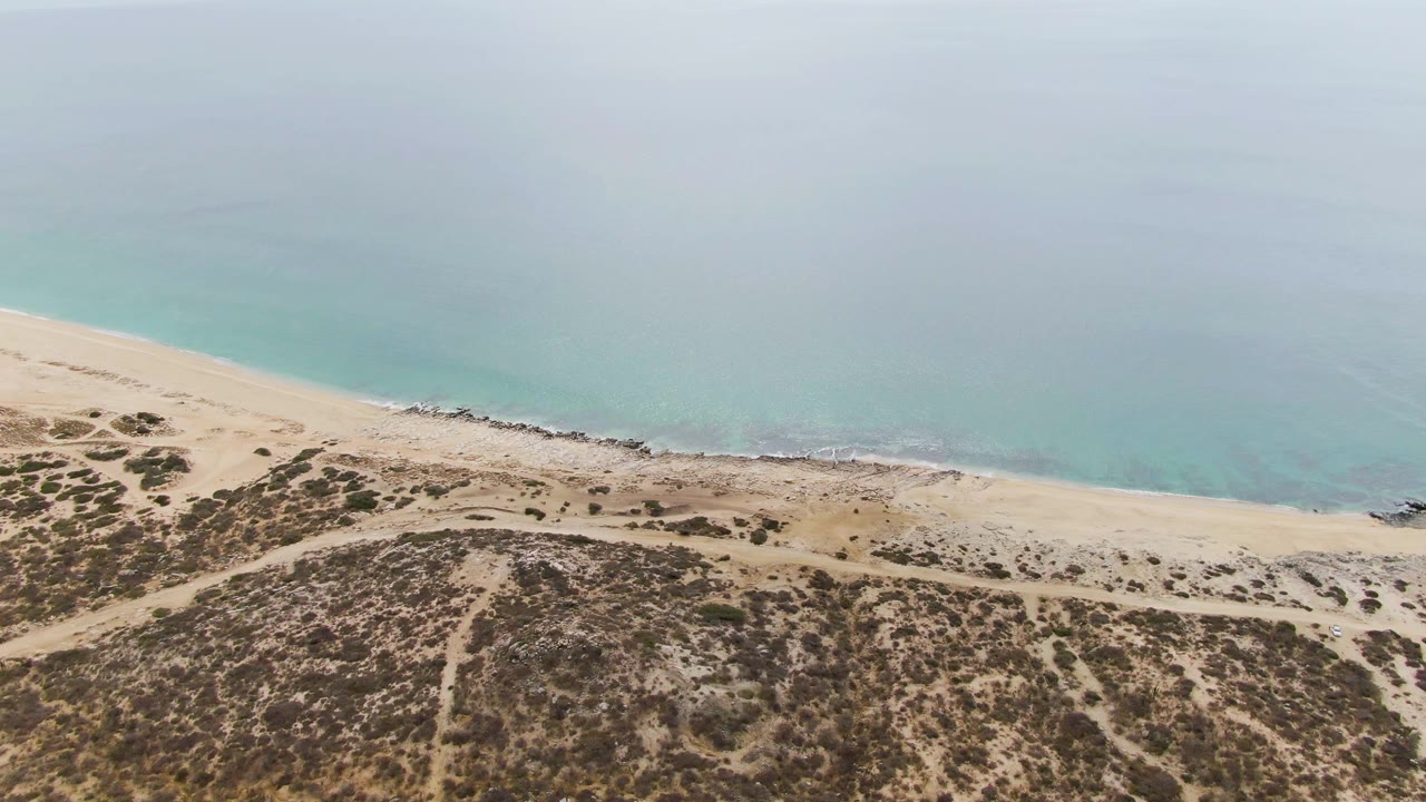 Aerial view at Punta Gorda Beach showing more of the sandy beach curve, low desert vegetation, faint paths through the dunes, turquoise ocean, and no visible people.