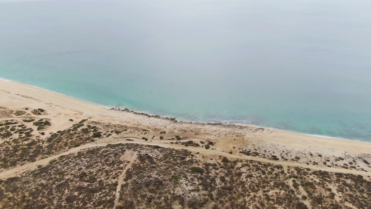 Aerial frame at Punta Gorda Beach with the shoreline shifted lower in view, scrub dunes in the foreground, pale sand across the middle, soft turquoise ocean above, and no visible people.