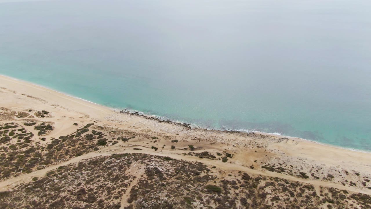 Opening aerial frame over Punta Gorda Beach, Baja California Sur, with scrub-covered dunes below, a pale sand strip, turquoise water above, and no visible people.