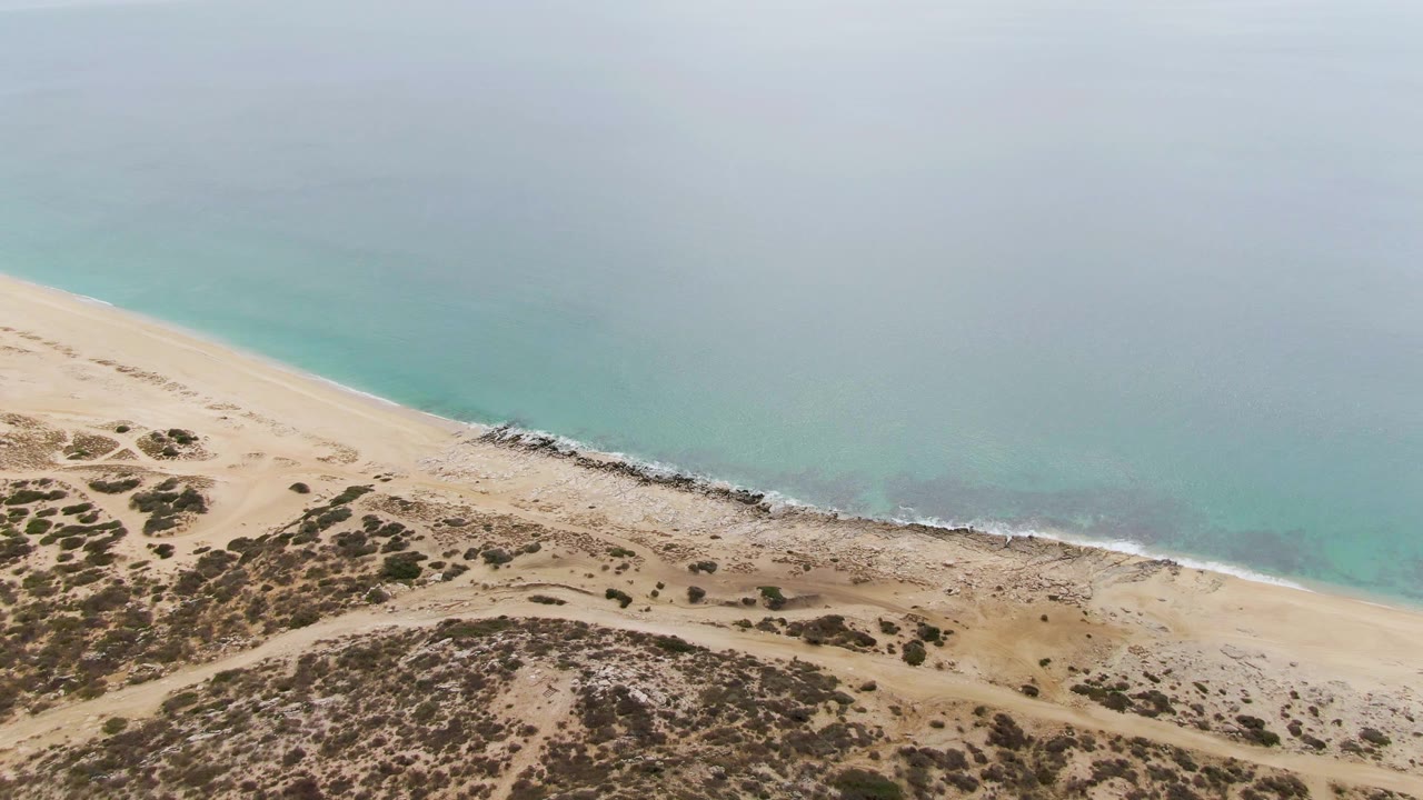 Mid-clip aerial view of Punta Gorda Beach with a broad band of sand, scattered dune vegetation, faint wave foam along the rocks, and no visible people.