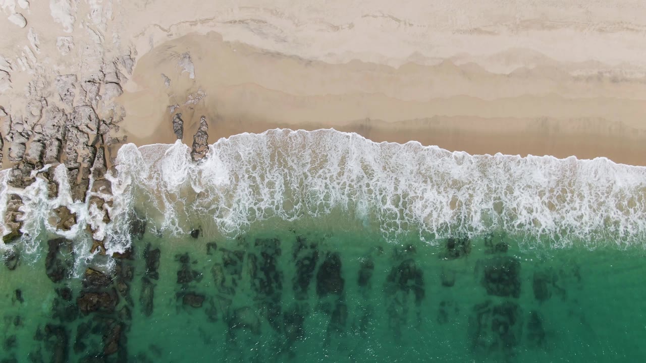 Overhead frame from Punta Gorda Beach, Baja California Sur, showing white surf spread across a rocky shoreline, beige sand above, turquoise water below, and no visible people.