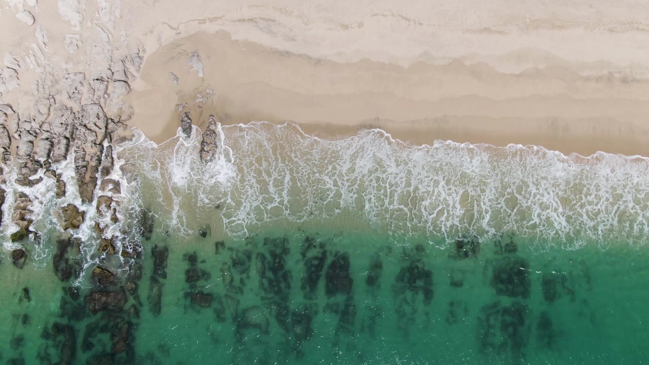 At 0:02, the overhead view shows surf foam stretched in a long horizontal band, with rocky outcrops interrupting the beach edge at Punta Gorda Beach and no visible people.
