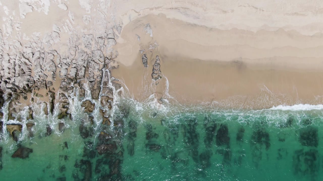 Overhead frame at Punta Gorda Beach, Mexico, with turquoise water below, white surf crossing dark rocks, pale sand above, and no visible people.
