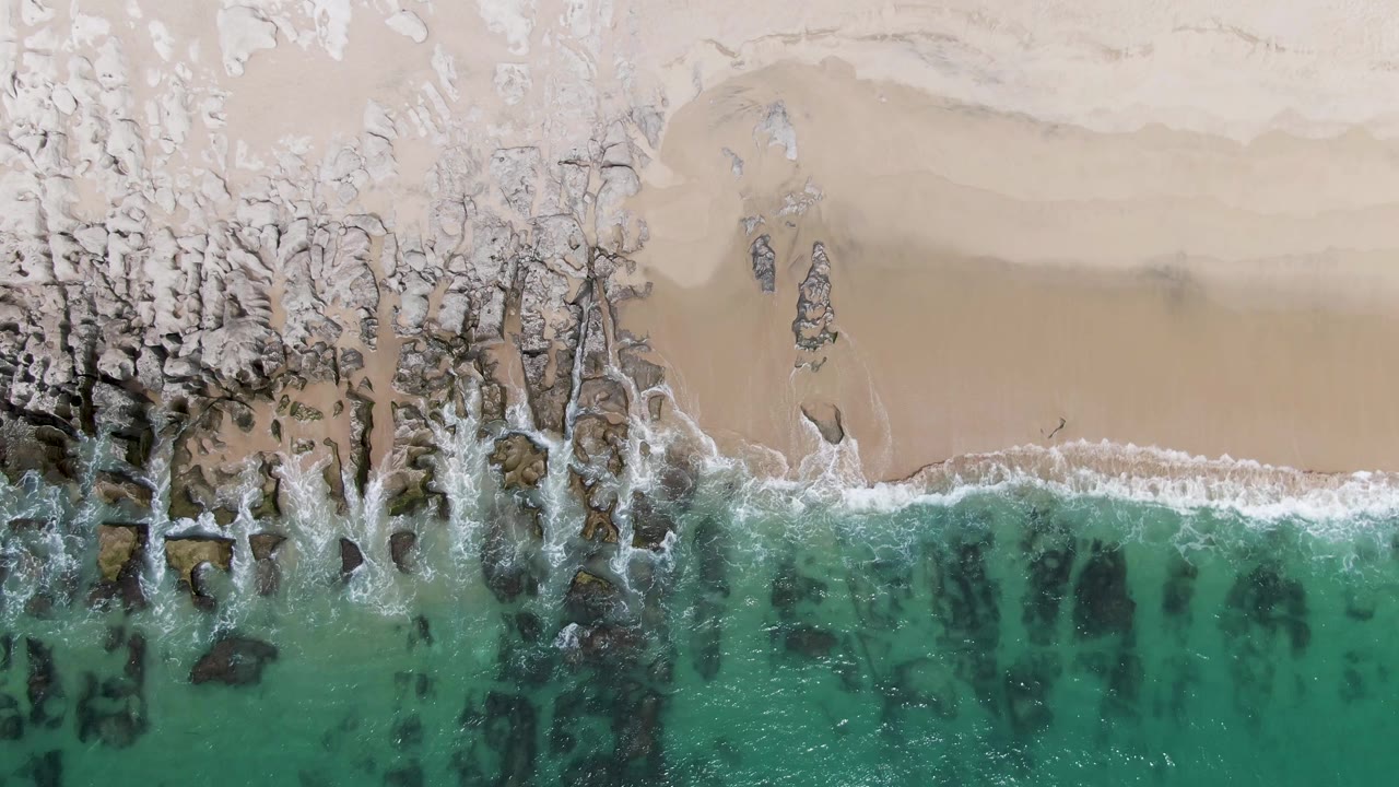 Aerial frame from Punta Gorda Beach showing green-blue water, broken white foam around dark rocks, pale sand, and no visible people.