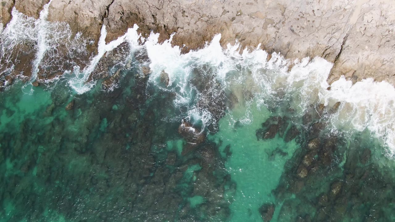 Aerial frame from Punta Gorda Beach showing white foam widening across dark submerged rocks beneath clear green-blue water, with no visible people.