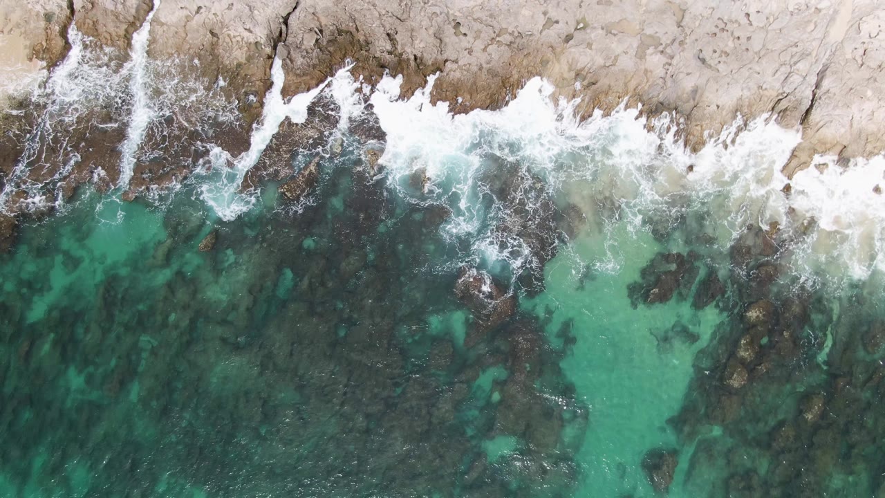 Top-down Punta Gorda Beach shoreline frame with foamy surf along the upper rocky edge and transparent turquoise shallows revealing scattered rocks, with no visible people.