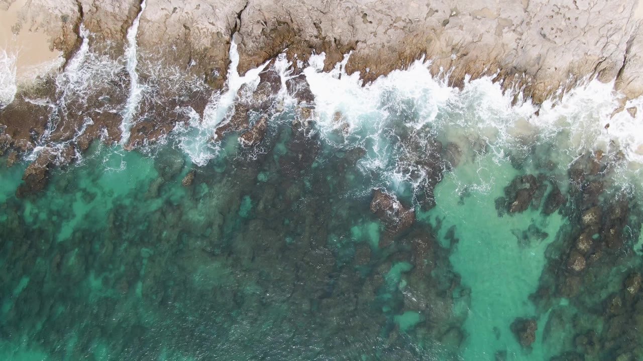 Overhead view at Punta Gorda Beach with a band of white surf crossing the rocky shore, darker water patches below, and no visible people.