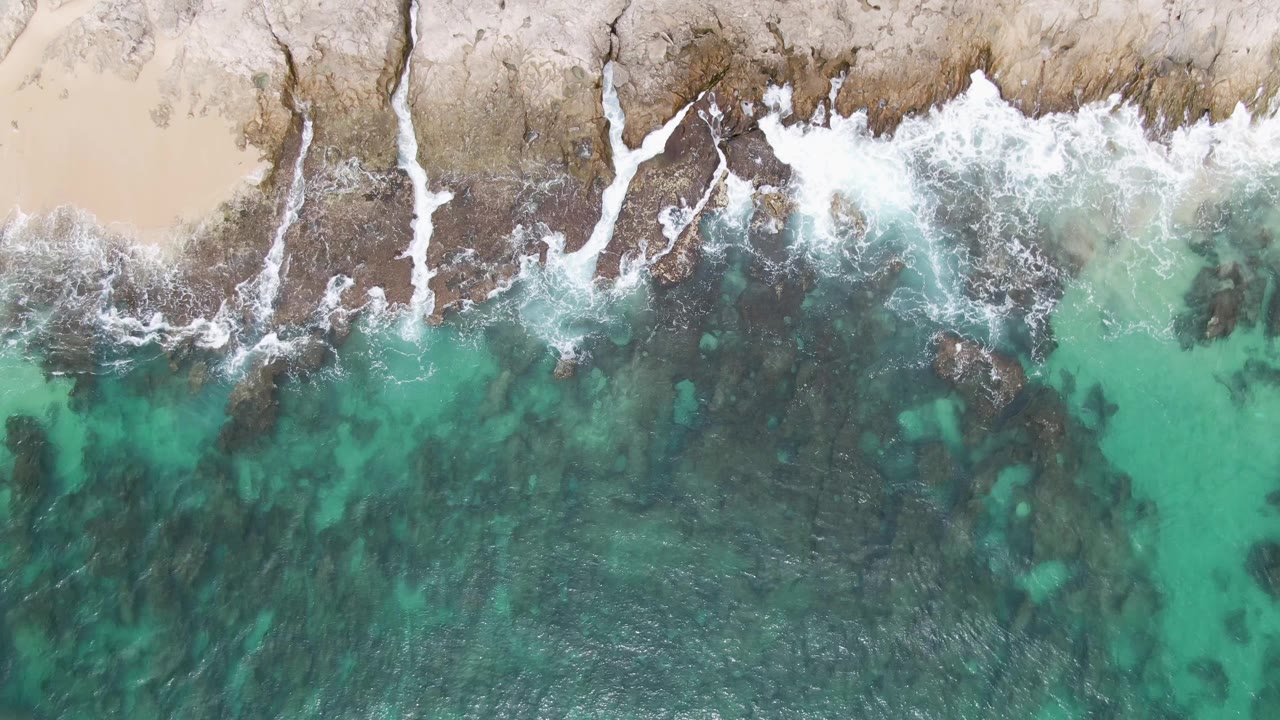 Overhead view of Punta Gorda Beach with white surf breaking along dark rocks, pale sand at the top edge, turquoise water below, and no visible people.