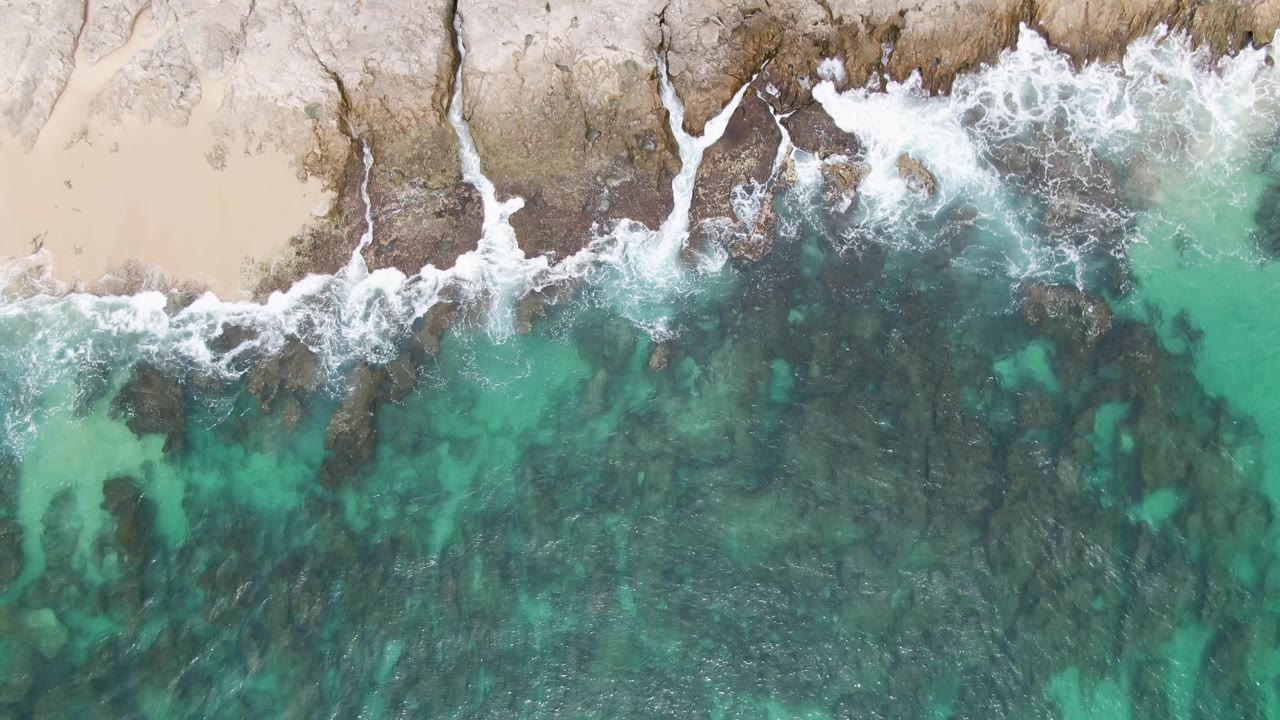 At 0:02, white surf curls over the Punta Gorda Beach rocks while darker underwater patches show through the turquoise shallows, with no visible people.