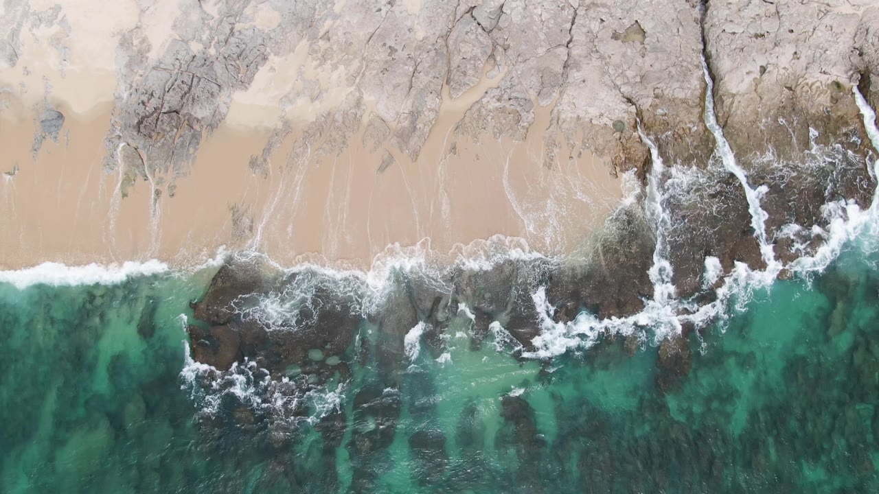 Top-down aerial frame of Punta Gorda Beach BCS, Mexico, with turquoise water and white foam reaching dark rocks beside pale sand, no visible people.