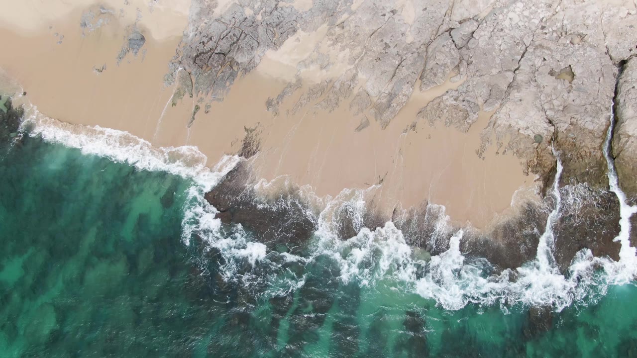 Top-down frame of Punta Gorda Beach BCS, Mexico, with a narrow white wave edge sliding along pale sand and dark rocks under green-blue water, no visible people.