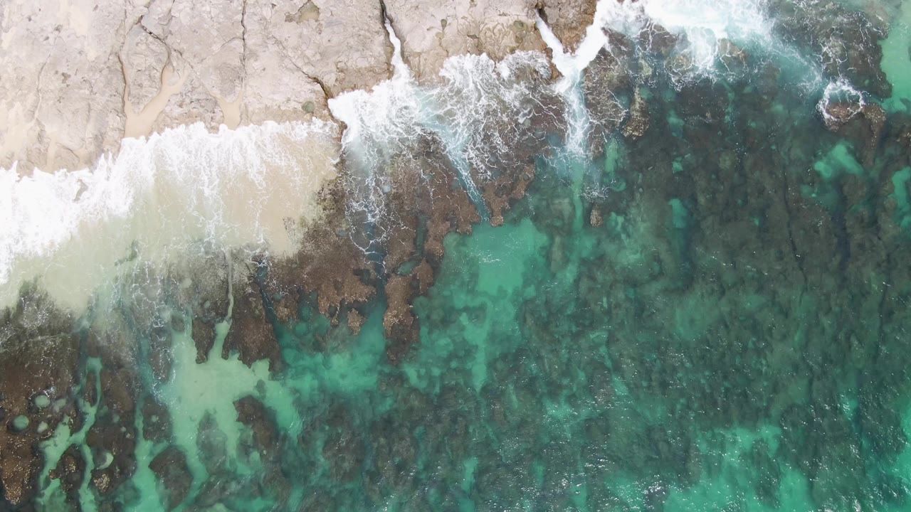 Overhead view at Punta Gorda Beach showing white surf washing over pale sand and dark coastal rocks beside clear turquoise water, with no visible people.