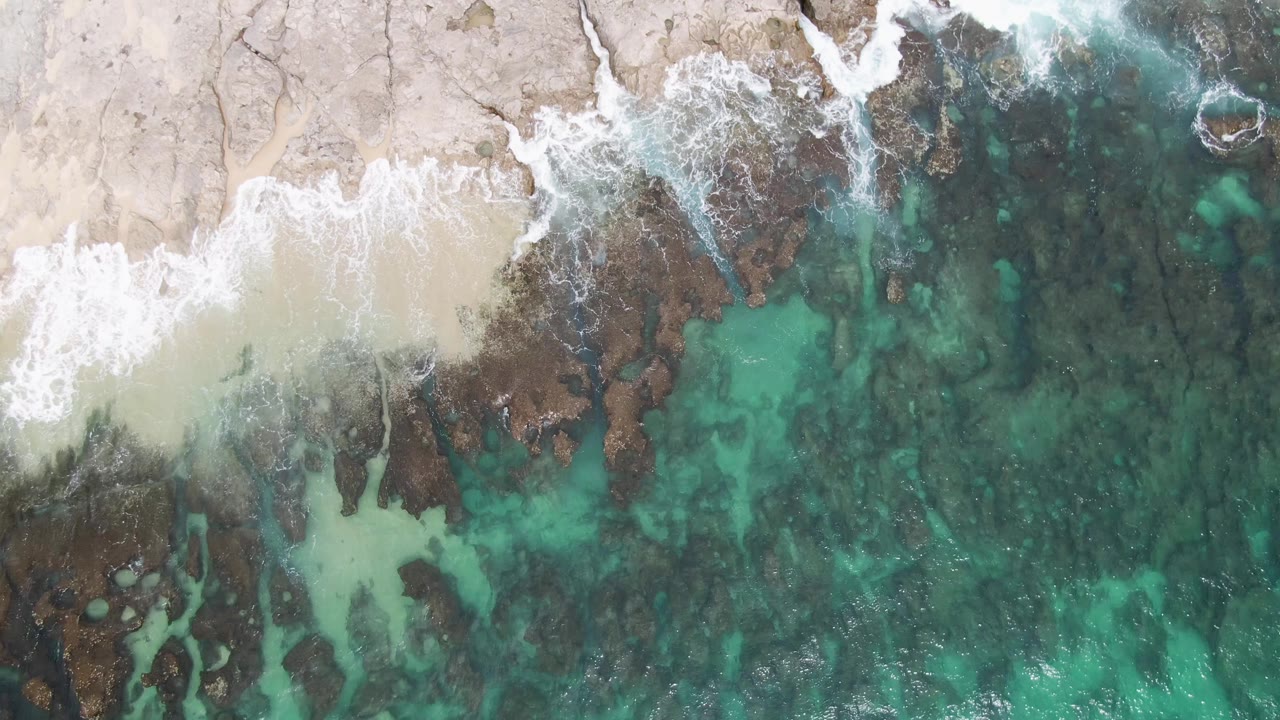 Aerial frame from Punta Gorda Beach with foam spreading across the rocky edge while green-blue water reveals darker seabed shapes, with no visible people.