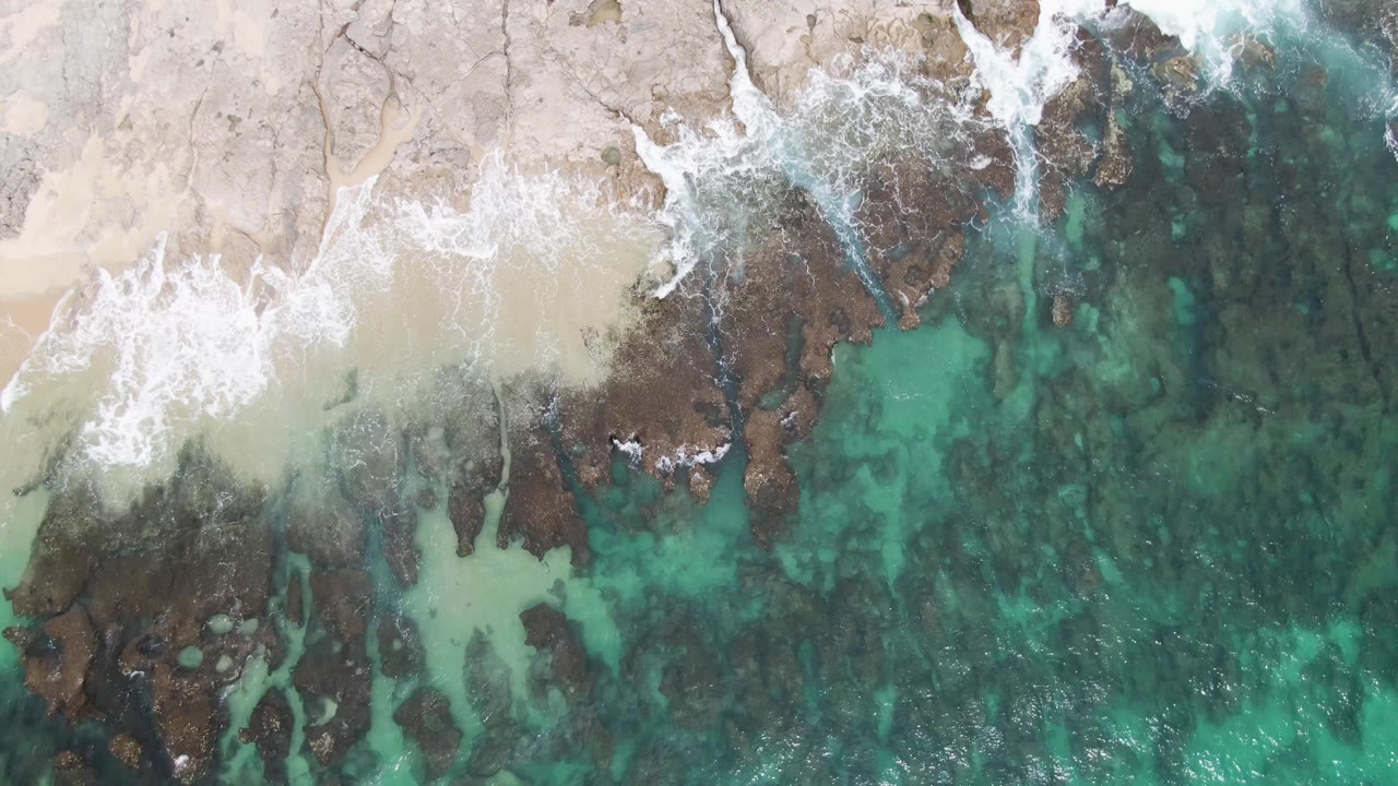 Top-down Punta Gorda Beach view where a wave curls around dark rocks between the sand and transparent turquoise water, with no visible people.