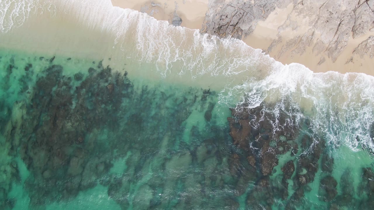 Aerial frame at Punta Gorda Beach showing foam pulling back from the sand while dark rock shapes remain visible through green-blue water, with no visible people.
