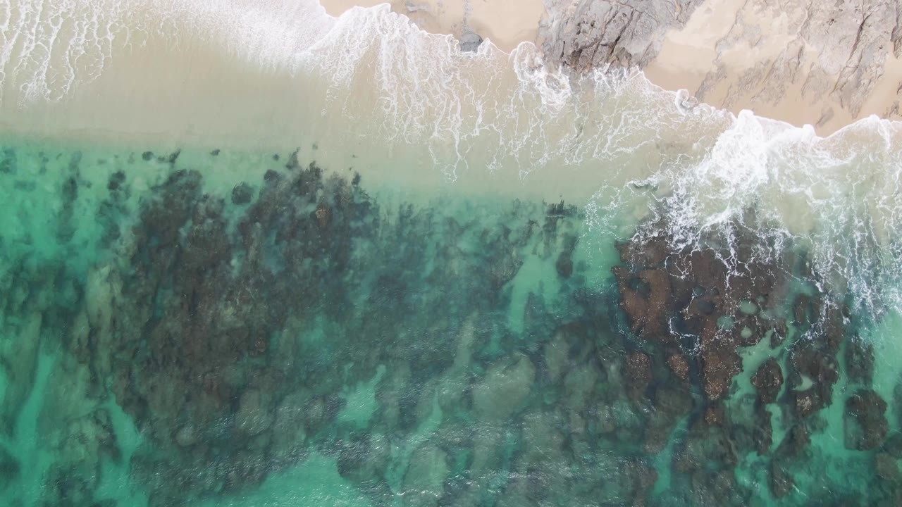 Top-down Punta Gorda Beach view with a bright band of surf curling along the sand and a cluster of dark rocks under turquoise water, with no visible people.
