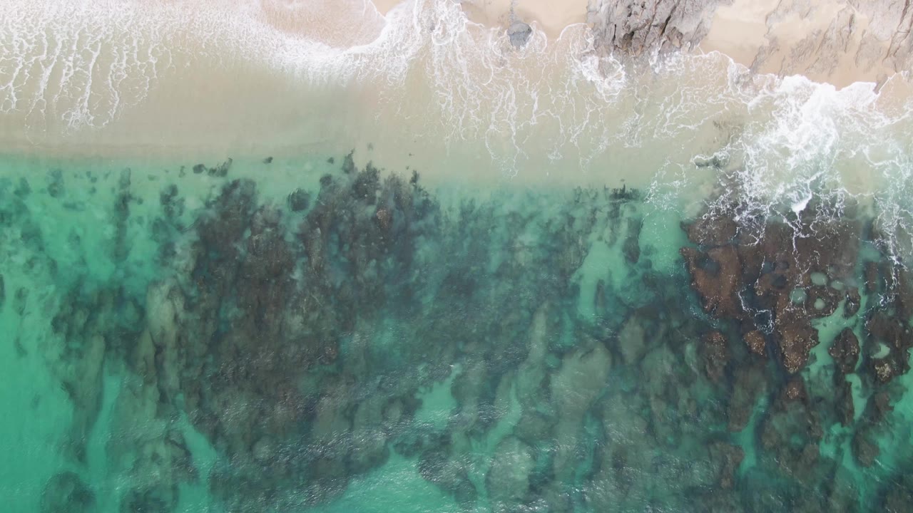Overhead frame from Punta Gorda Beach where the sand runs across the top, white foam breaks around a central rock patch, and clear turquoise water fills the lower frame, with no visible people.