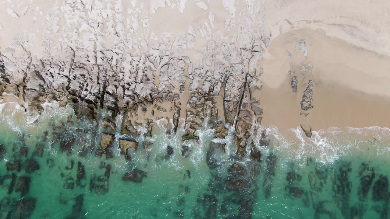Aerial frame of Punta Gorda Beach with turquoise water pushing white foam over dark shoreline rocks, pale sand above, and no visible people.