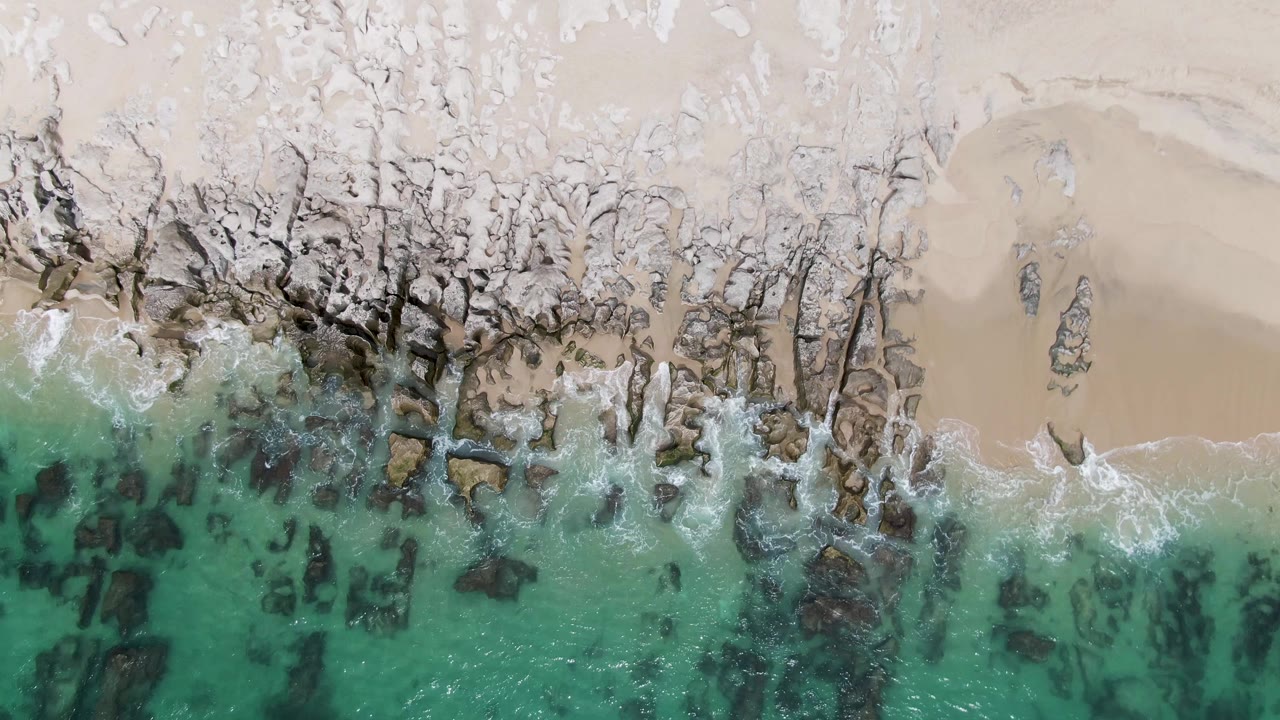 Aerial frame at Punta Gorda Beach where a thin white wave line spreads across jagged rocks beneath a smooth sand section, with no visible people.