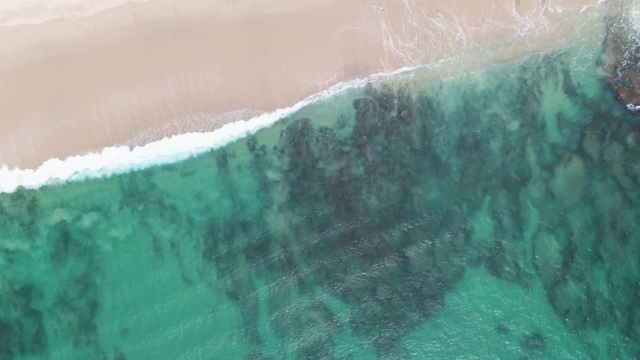 Aerial view at Punta Gorda Beach with a diagonal foam edge sliding along pale sand and transparent green-blue water over darker seabed patches, with no visible people.
