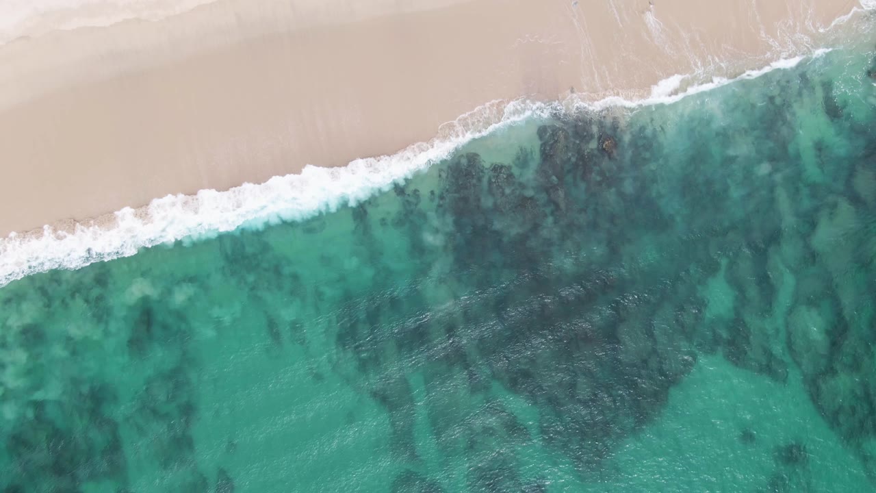 Overhead Punta Gorda Beach frame showing a thicker band of white breaking surf between the sandy upper edge and turquoise shallows with dark underwater rocks, with no visible people.