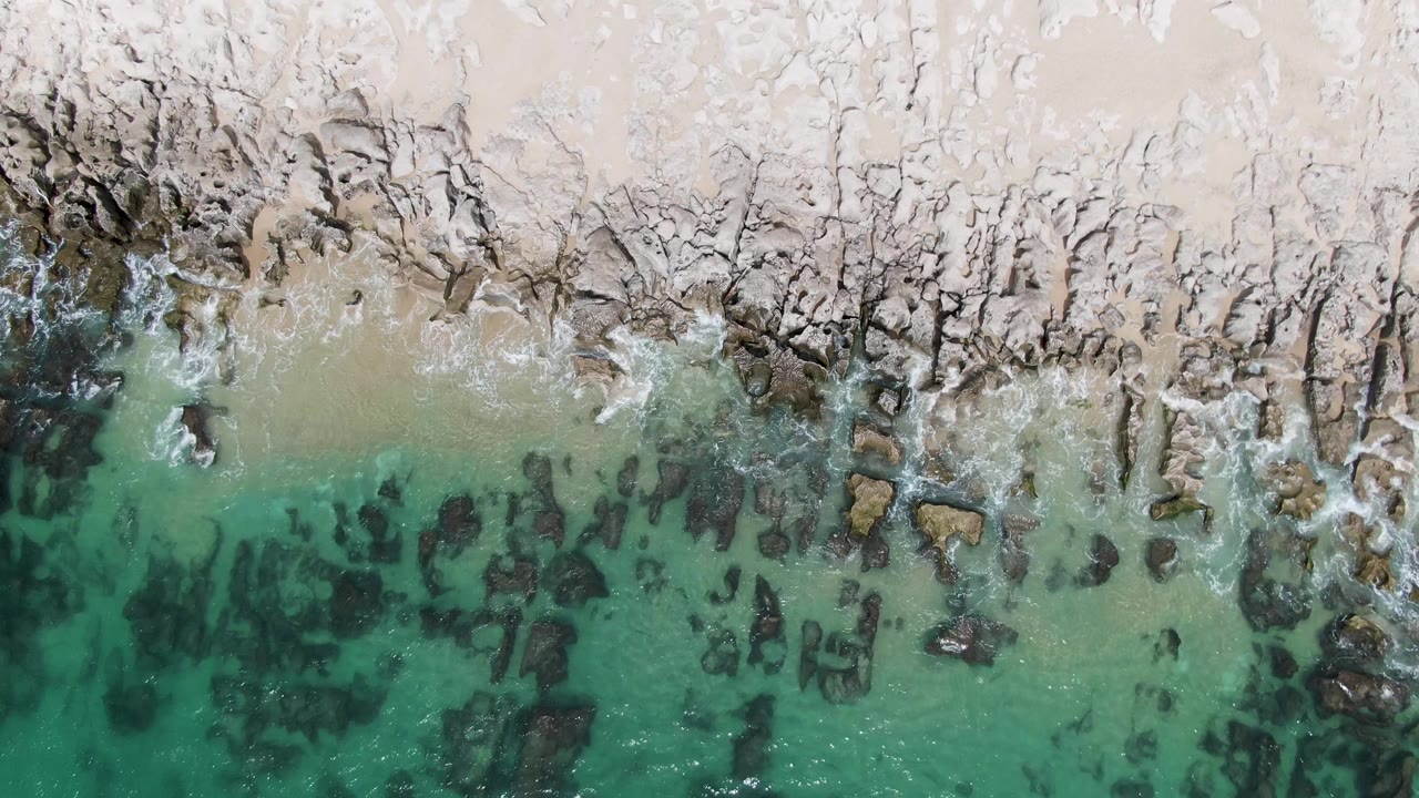 Overhead frame of Punta Gorda Beach with a denser cluster of dark submerged rocks in the lower water, pale sand across the top, light foam on the shoreline, and no visible people.
