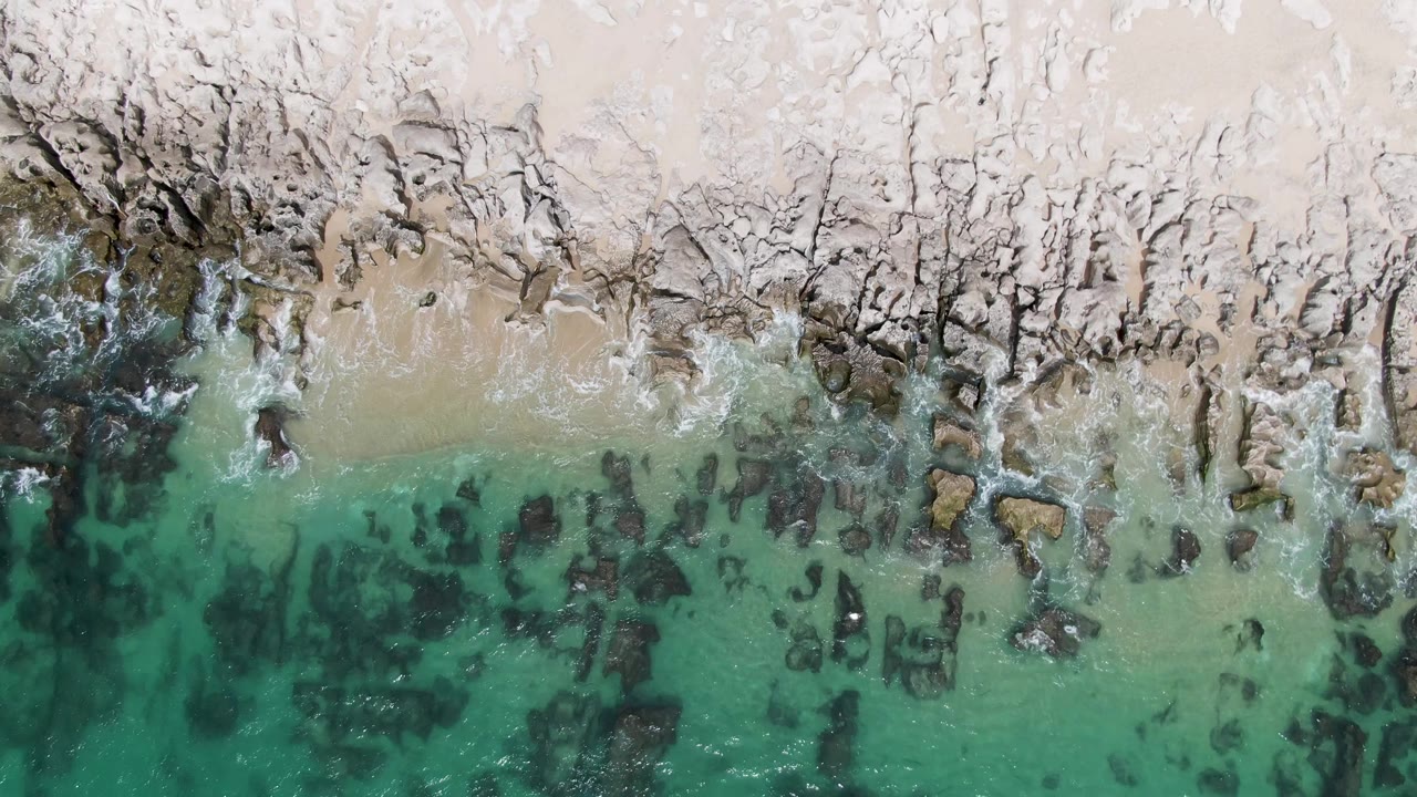 Overhead frame of Punta Gorda Beach where small white waves break around exposed rocks, with sandy beach above and green-blue water below, and no visible people.