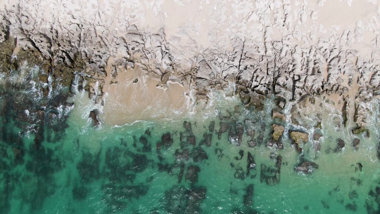 Overhead frame of Punta Gorda Beach with a darker water channel at left, scattered rocks in the shallows, pale sand at top, and no visible people.