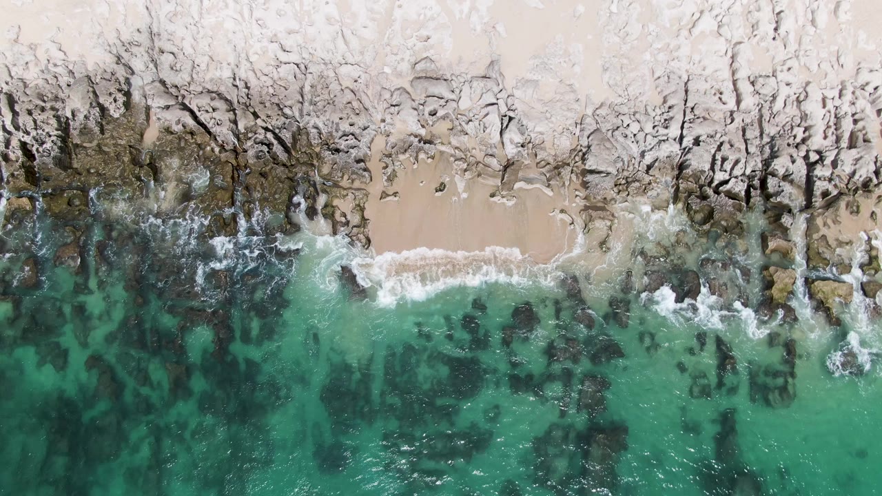 Overhead turquoise water meeting sandy beach and dark rocks