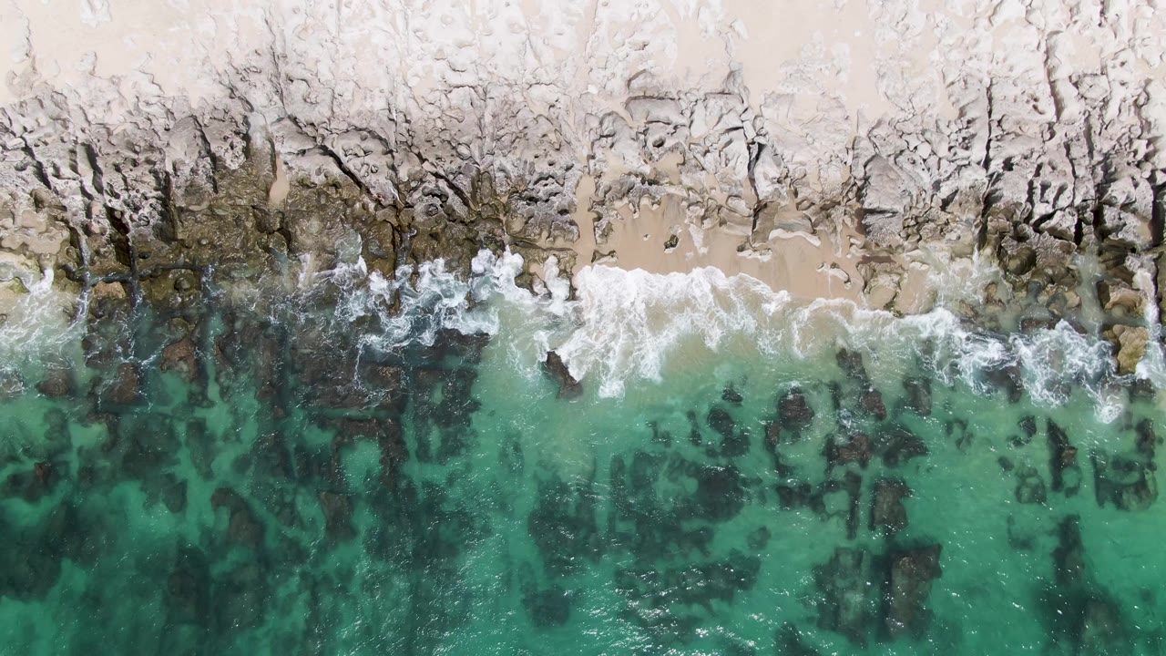 At 0:02, a broader band of white surf breaks along the pale sand and dark rocks at Punta Gorda Beach, with turquoise water filling the lower frame and no visible people.