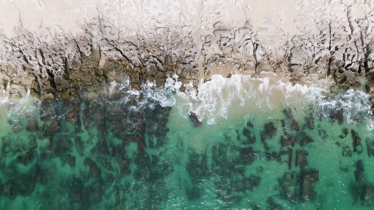 At 0:03, foamy wash thins across the rocks and sand at Punta Gorda Beach while darker underwater rock shapes remain visible in the turquoise water, with no visible people.
