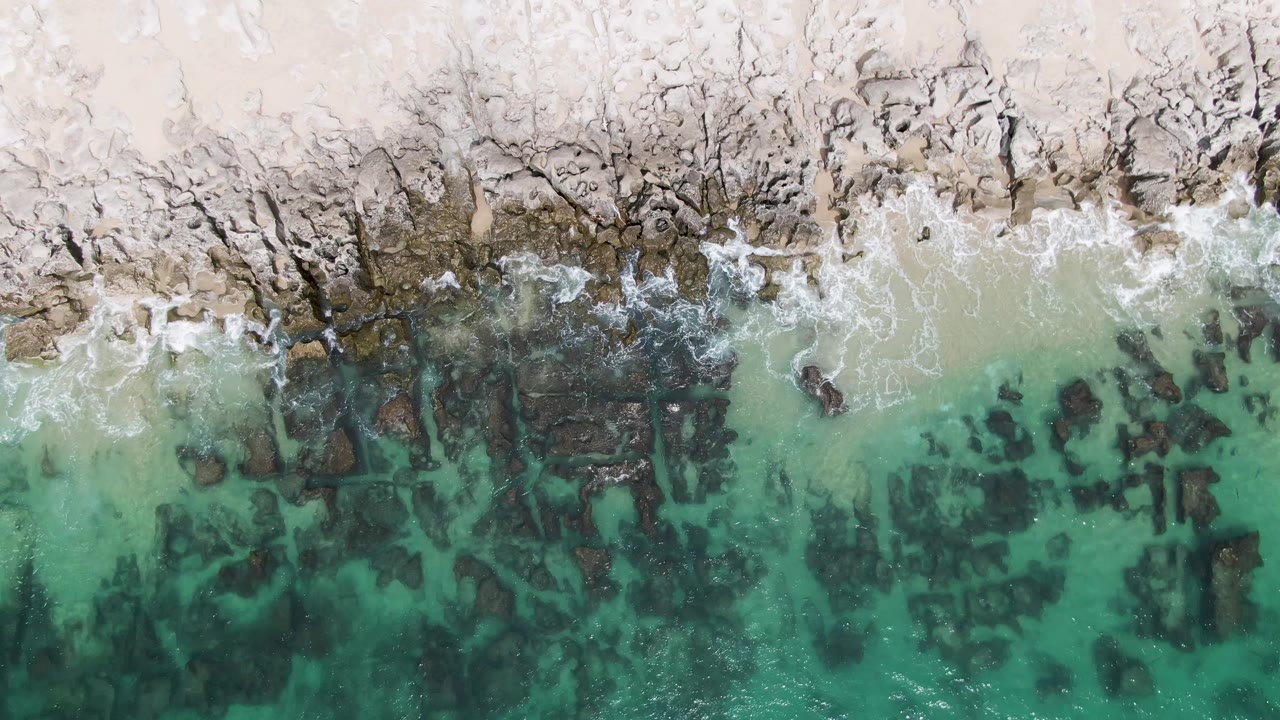 Overhead frame from Punta Gorda Beach BCS showing pale sand, jagged dark rocks, turquoise shallow water, scattered foam, and no visible people.