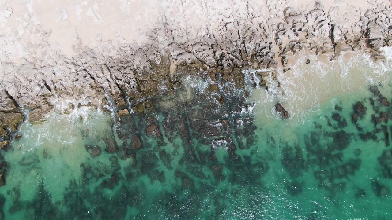 At 0:02, the overhead Punta Gorda Beach BCS view holds sand at the top, broken rocks through the center, darker submerged shapes, turquoise water, and no visible people.