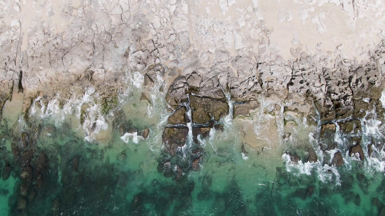 Overhead frame at Punta Gorda Beach BCS showing broken rock shelves, pale sand above them, and white surf lines shifting along the clear green water, with no visible people.