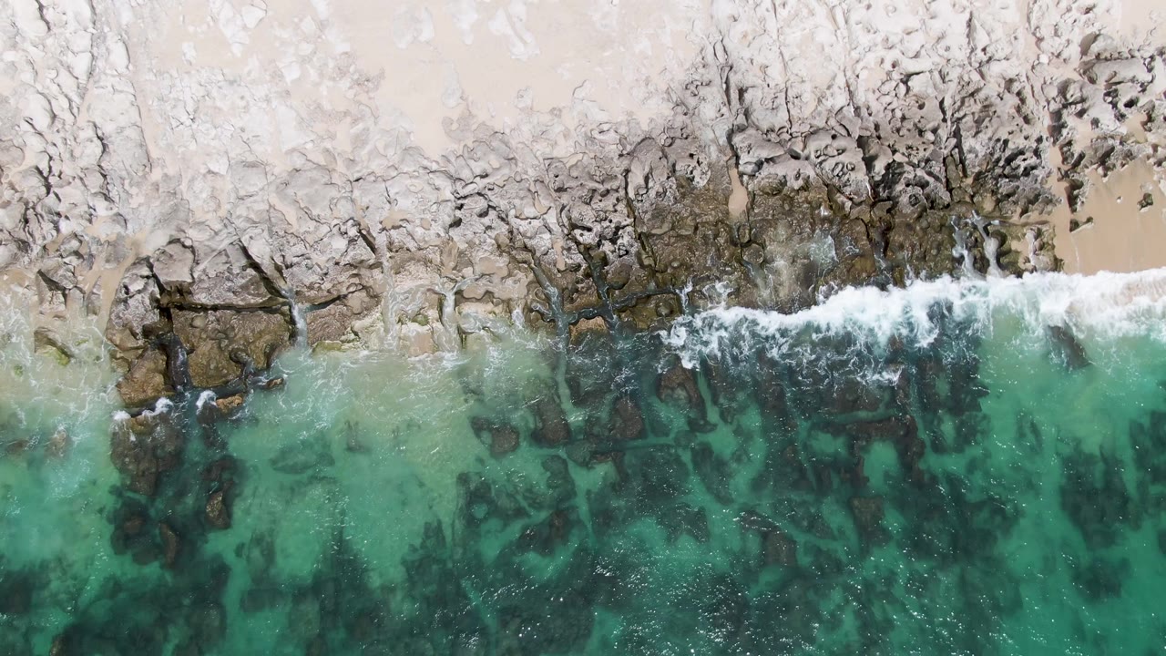 Overhead view of Punta Gorda Beach in Baja California Sur, with clear turquoise water over dark rocks and a narrow band of white surf along the pale shore, no visible people.