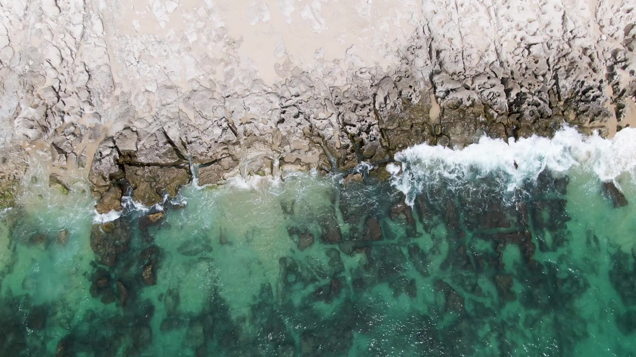 A later overhead frame at Punta Gorda Beach shows a broader patch of white surf breaking over shoreline rocks, with green-blue water revealing the darker seabed below and no visible people.
