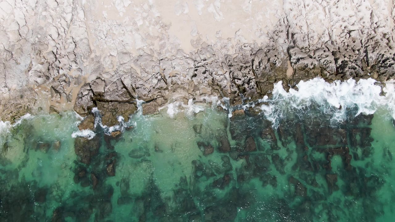 Over Punta Gorda Beach, the foam line sits tight against the rocky coast while translucent turquoise water fills the lower frame with visible dark rock patches and no visible people.