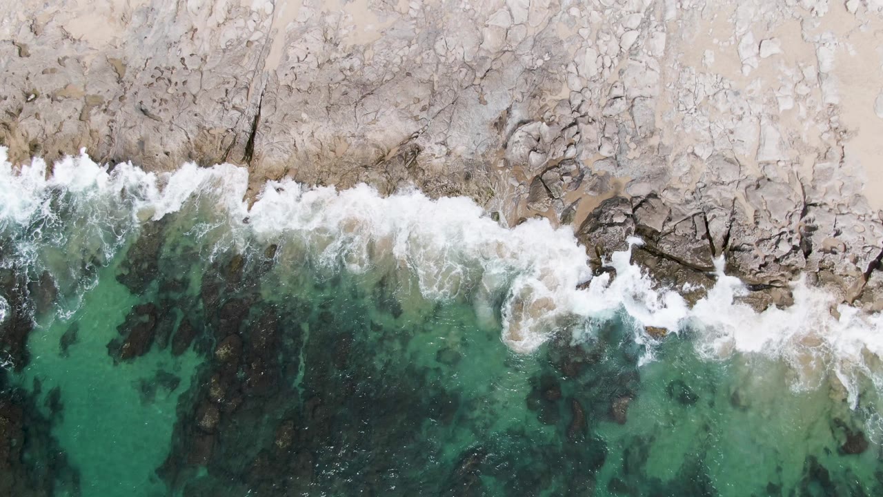 At Punta Gorda Beach, white surf curls across dark submerged rocks below a pale rocky coast, with green-blue water filling the lower frame and no visible people.