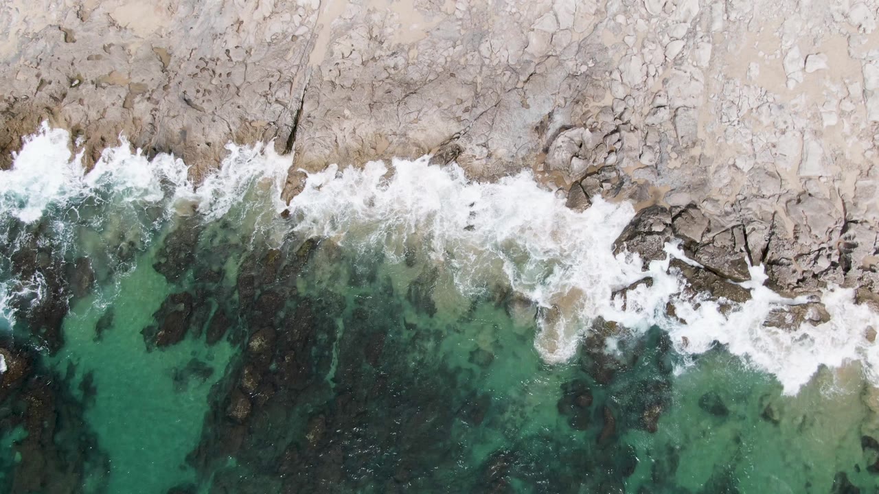 A top-down frame from Punta Gorda Beach shows foam spreading unevenly across the rocky coastal edge, with turquoise shallows below and no visible people.