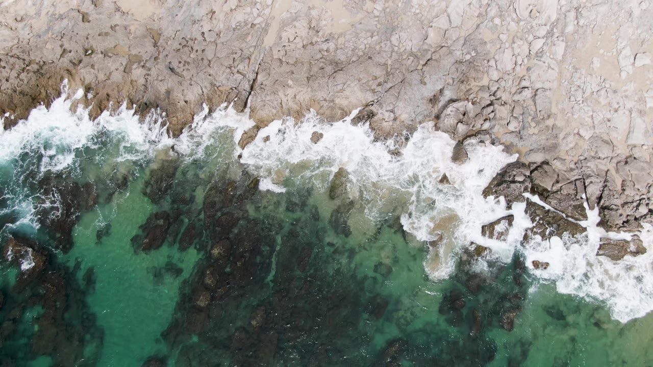 Over Punta Gorda Beach, foamy water breaks across angular shoreline rocks while clear green water and darker rock patches sit below, with no visible people.