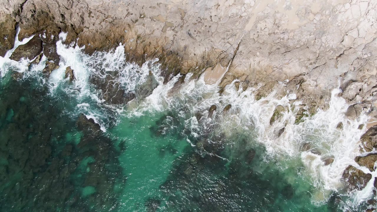 Top-down coastal view at Punta Gorda Beach showing white surf wrapping around exposed rocks, with green water below the shoreline and no visible people.