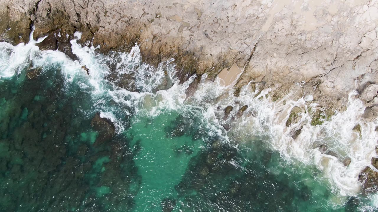Overhead drone-style view of a foamy wave breaking along the rocky Punta Gorda Beach shoreline, with turquoise water below and no visible people.