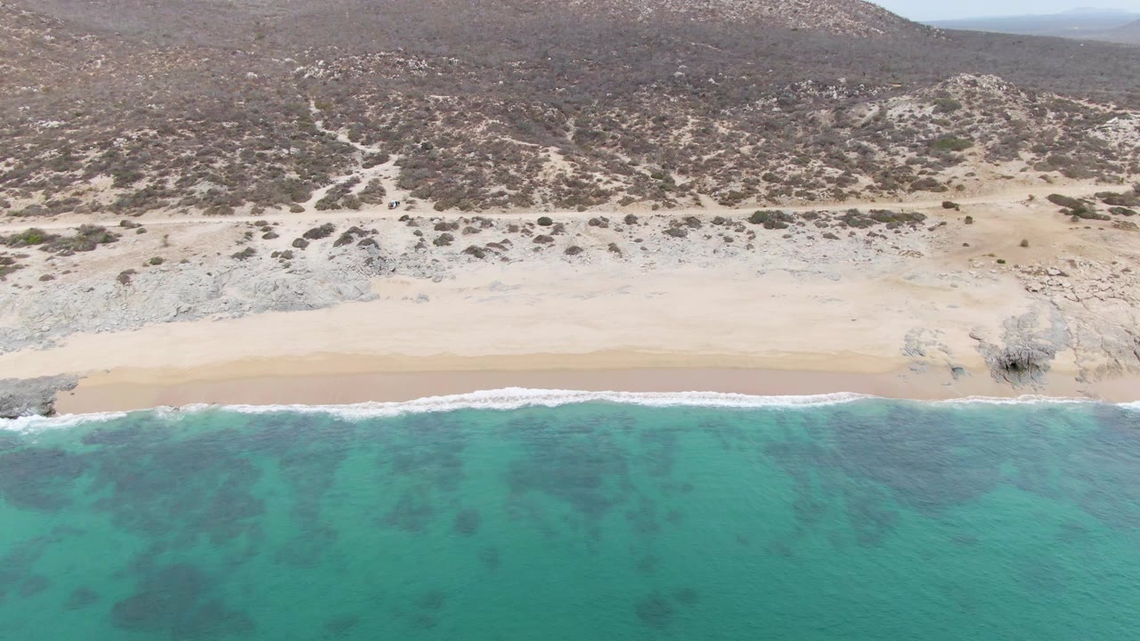 Aerial view of turquoise ocean meeting a sandy beach below a dry rocky hillside.