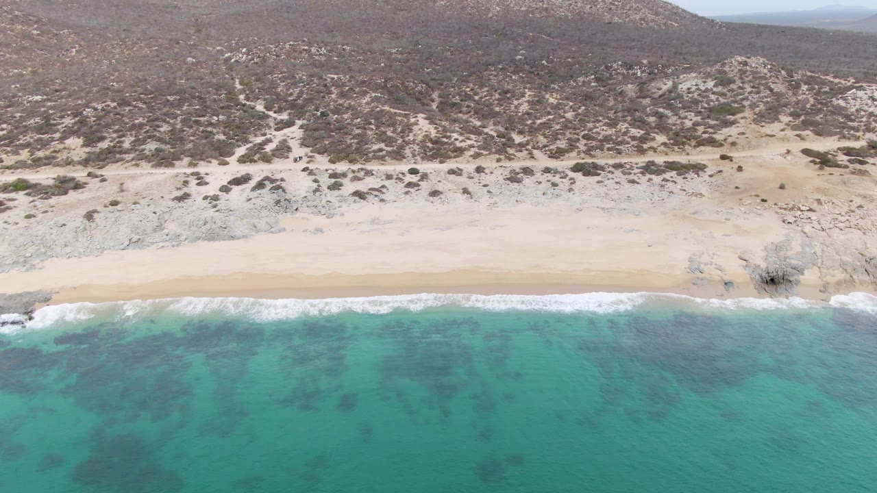At 0:02, shallow turquoise water shows darker patches near the surf, with a smooth sandy strip and rocky Baja California Sur hillside behind it, with no visible people.