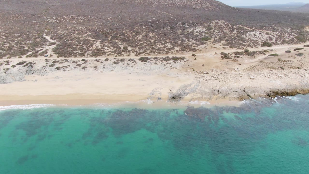 Aerial view at Punta Gorda Beach in Baja California Sur with turquoise water in front, a pale sand strip, rocky shoreline, scrub-covered hills, and no visible people.