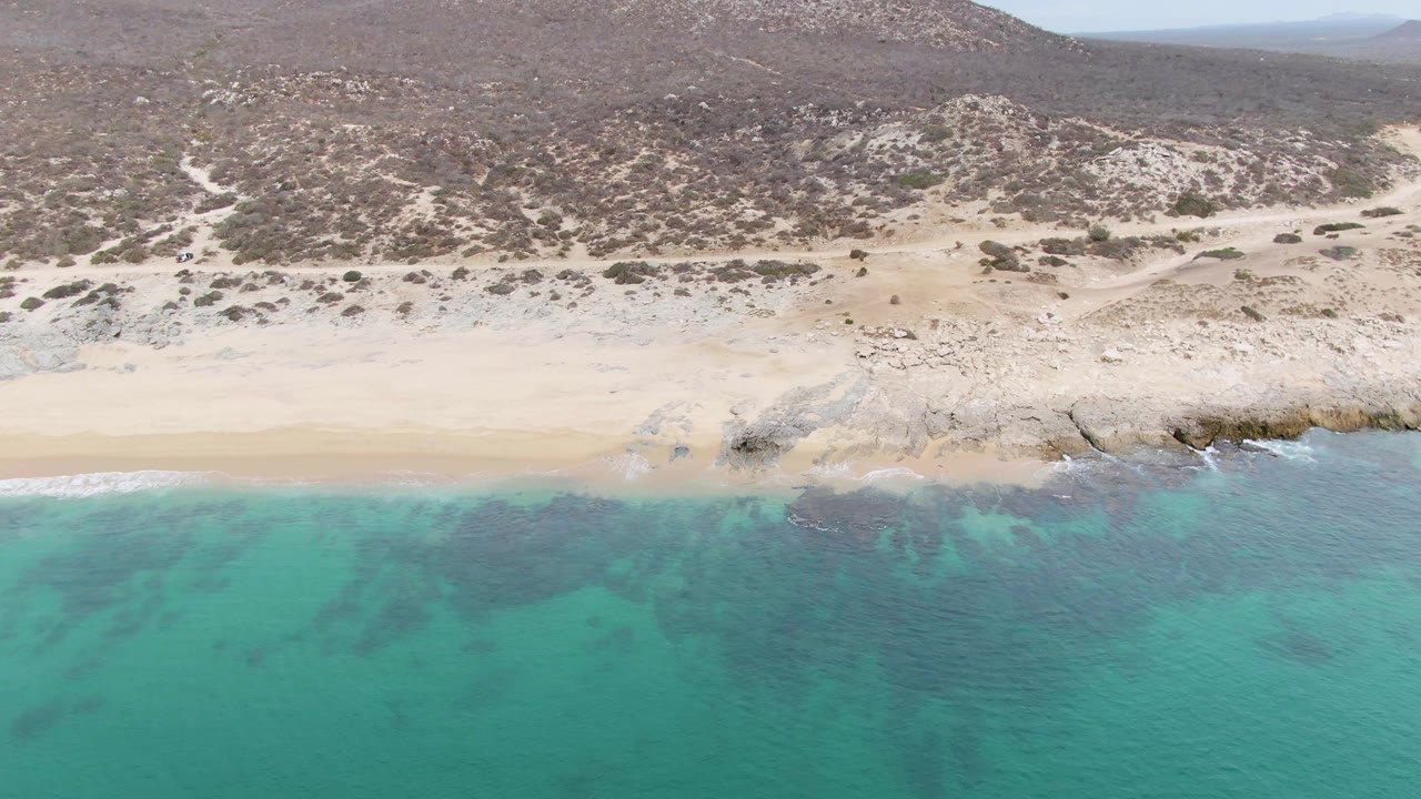 Drone view over blue-green water toward Punta Gorda Beach, with light surf touching the sand, rocky outcrops near shore, low desert hills behind, and no visible people.