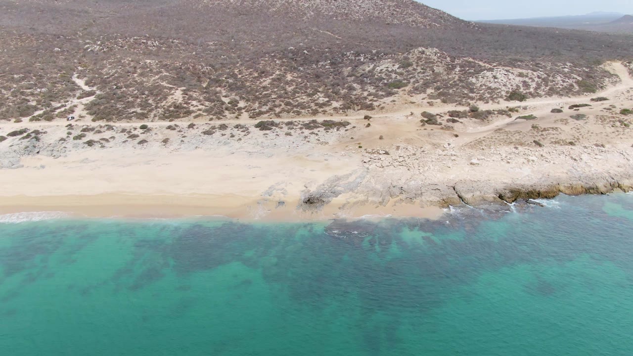 Wide aerial frame of Punta Gorda Beach showing turquoise ocean across the lower half, pale sand and rocks at the shoreline, brown scrubland hills beyond, and no visible people.