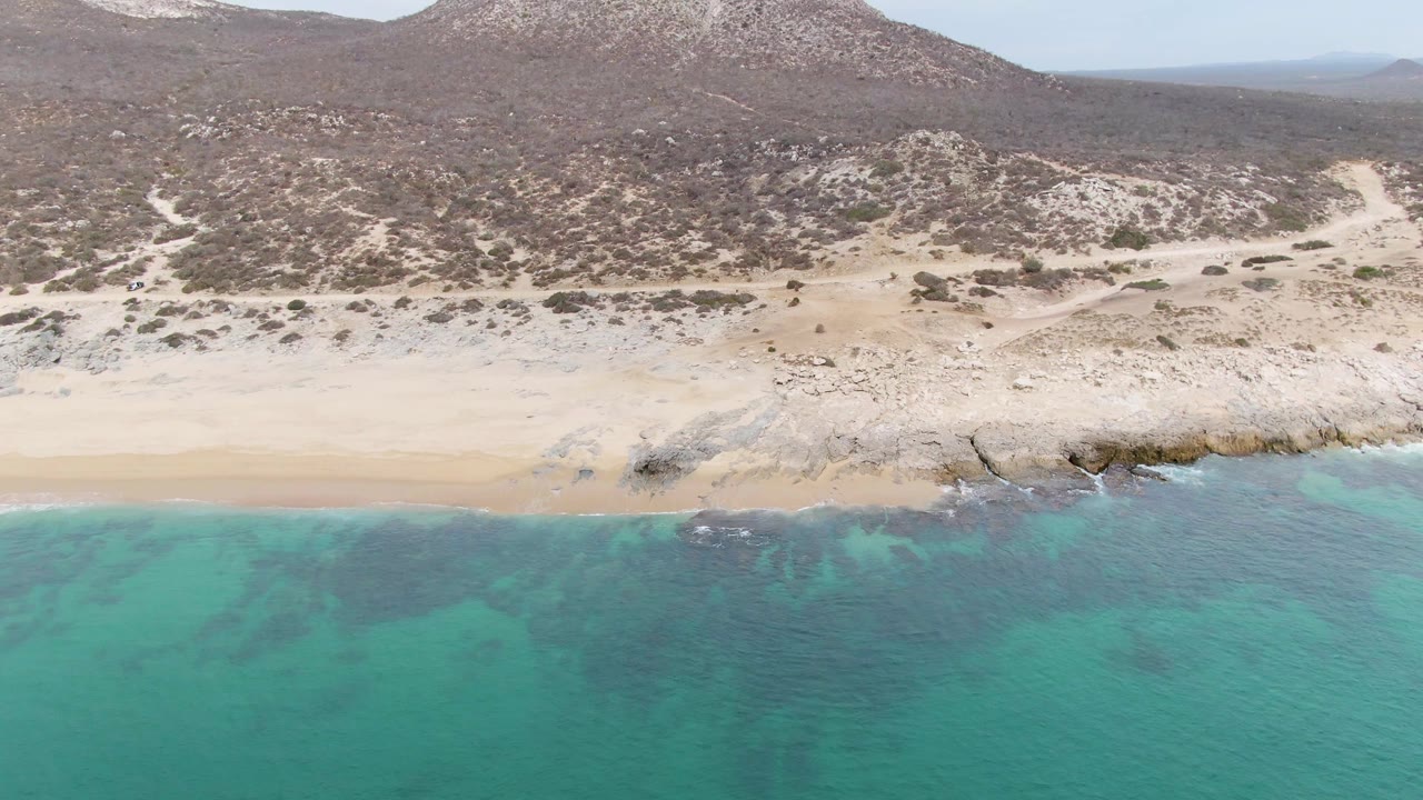 Aerial view at Punta Gorda Beach with a rocky shoreline feature near center, sand to the left, turquoise water below, desert vegetation on the hills, and no visible people.